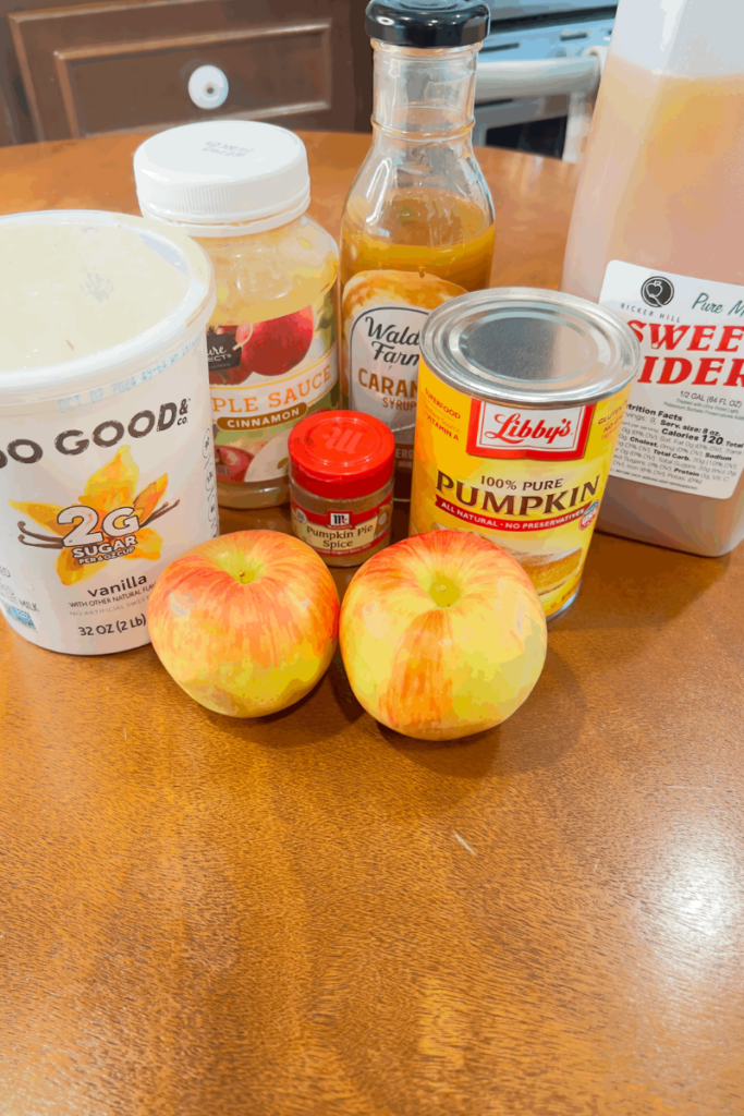 ingredients lined up on the table needed for apple pumpkin smoothie to include containers of yogurt, applesauce, caramel sauce, pumpkin puree can, 1/2 gallon of apple cider and pumpkin pie spice bottle