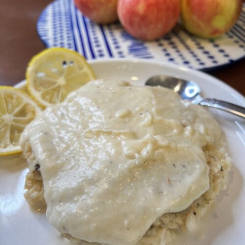 white plate with pureed shrimp Alfredo dish, two slices lemon on the plate with a spoon and a pile of apples in the background