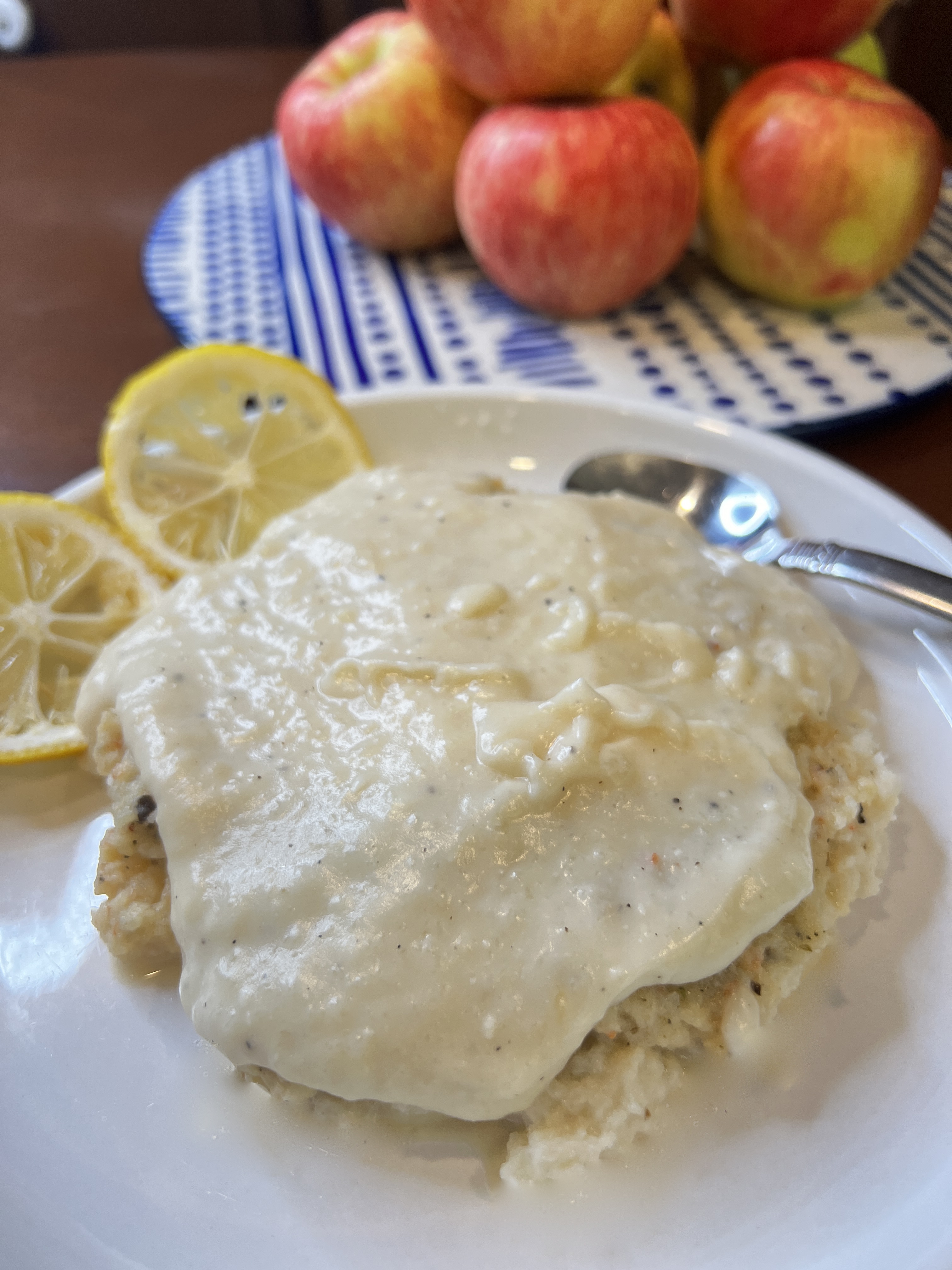 white plate with pureed shrimp Alfredo dish, two slices lemon on the plate with a spoon and a pile of apples in the background