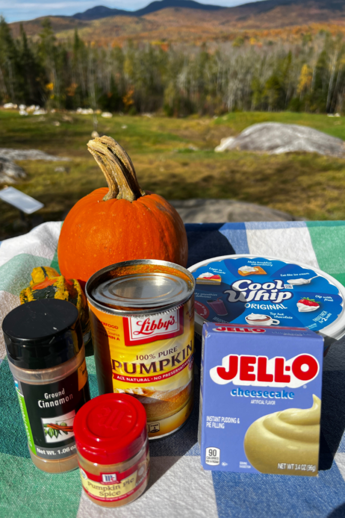 tabletop  with a small pumpkin and ingredients for a recipe to include spice jars of cinnamon and pumpkin pie spice, can of pumpkin puree, box of jello pudding mix and container of cool whip
