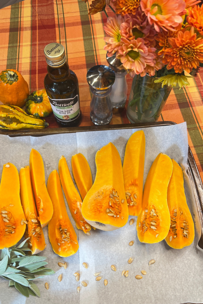 baking sheet of butternut squash chunks with bundle of fresh sage leaves. on the table ornamental pumpkins, bottle of olive oil, salt and pepper mills and vase of fall flowers