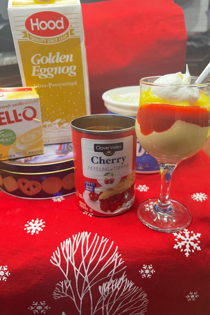 Line up of all ingredients needed to make a Christmas dessert including jello box, eggnog, tin of shortbread cookies, cherry pie filling and cool whip